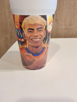 McDonald's pappersmugg med fotbollstema - Säljer en färgglad pappersmugg från McDonald's med fotbollstema och tecknade illustrationer av spelare och texten 'Winners Yame'. Muggen har en vit topp och motiv i orange, blått och gult. Perfekt för samlare eller som dekoration.