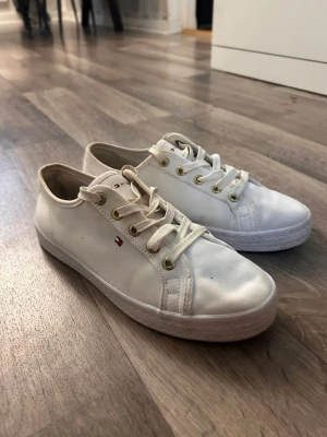 Vita låga sneakers Tommy Hilfiger - Snygga vita sneakers från Tommy Hilfiger med guldfärgade öljetter och klassisk logga på sidan. Skorna har lågt skaft, vita snören och ovandel i textil. Perfekta för en clean och stilren look.