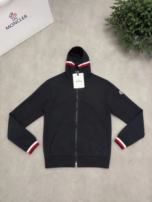 Svart Moncler hoodie med dragkedja - Snygg svart hoodie från Moncler med hel dragkedja, huva och fickor framtill. Rödvita ränder på huva och ärmslut ger en sportig vibe. Moncler-logga på ärmen. Perfekt för dig som vill ha en stilren och exklusiv look.