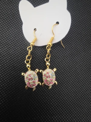 Handgjorda Guldiga örhängen med sköldpaddor - Snygga handgjorda hängande örhängen i guldfärg med sköldpaddsformade hängen. Sköldpaddorna har ett mönster med små rosa och gröna blommor på vit bakgrund. Perfekta för dig som gillar unika accessoarer med djurmotiv.