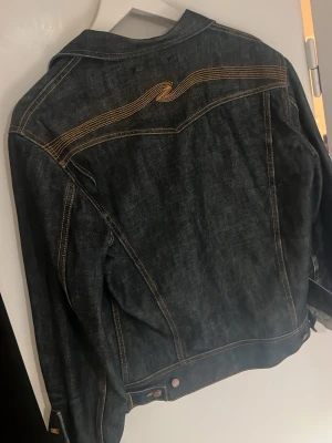 Vintage Nudie Conny Raw Denim Jacket - Snygg mörkblå jeansjacka från Nudie Jeans. Jackan har kontrastsömmar, två bröstfickor med knapp och en unik broderad Nudie Jeans detalj på ryggen, även riktigt snygga fades. Perfekt för dig som gillar streetwear och tidlös denimstil. Storlek M
