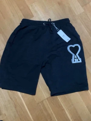 Svarta shorts från Ami Alexandre Mattiussi - Svarta shorts från Ami Alexandre Mattiussi med snörning i midjan och ett stort vitt hjärta med bokstaven A broderat på ena benet. Sköna och stilrena, perfekta för sommaren. Tillverkade i mjuk bomull.