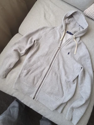 Grå hoodie från Polo Ralph Lauren - Snygg ljusgrå hoodie från Polo Ralph Lauren med dragkedja. Säljer denna snygga och bekväma zip-up fleece hoodie från Ralph Lauren på grund av viktökning. Hoodien är i toppskick och har mest legat i garderoben...                                            Skriv gärna vid frågor eller intresse!