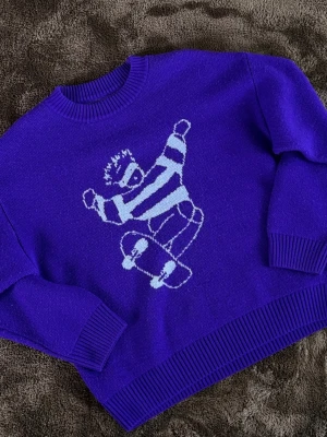 Lila stickad tröja från Polarskate - Cool lila stickad tröja från Polarskate med ett stort motiv av en skateboardåkare i vit på framsidan. Tröjan har rund halsringning och ribbade muddar vid ärmslut och nederkant. Perfekt för dig som gillar streetwear och unika prints. M fits S
