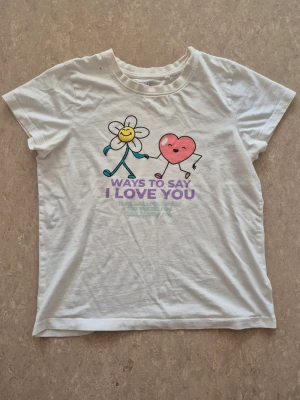 T-shirt - Vit t-shirt med ett färgglatt tryck av en glad blomma och ett hjärta som håller händer, samt texten 'Ways to Say I Love You' i lila. T-shirten har rund hals och korta ärmar, perfekt för dig som gillar lekfulla motiv. Finns små fläckar.