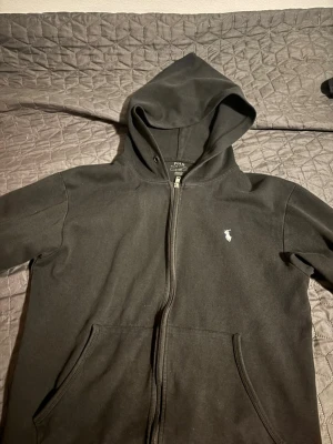 Svart zip hoodie från Polo Ralph Lauren - Säljer en svart hoodie med dragkedja från Polo Ralph Lauren. Klassisk design med huva, två fickor framtill och den ikoniska vita logon broderad på bröstet. Tillverkad i mjukt bomullsmaterial som är perfekt för chill dagar.