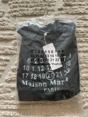 Maison Margiela svart t-shirt - Snygg svart t-shirt från Maison Margiela med det ikoniska numrerade trycket framtill och texten 'Maison Margiela PARIS'. Klassisk passform och rund hals. Tillverkad i mjuk bomull, perfekt till jeans eller shorts.