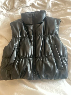 Svart pufferväst i fejkläder - Snygg svart pufferväst i fejkläder med hög krage och dragkedja framtill. Västen har två fickor på sidorna och en cool, puffig design med resårdetaljer. Perfekt för att lyfta din outfit med en trendig touch.