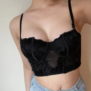Svart spetskorsett croppad topp - Säljer en svart korsettopp med spetsdetaljer. Toppen har bygel, croppad passform och tunna axelband. Perfekt för dig som vill ha en edgy och trendig look. 