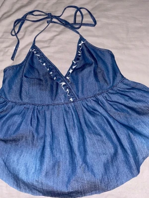 Blå denim linne topp med volang - blå denim halter linne/topp med knyt i nacken och v-ringning. Toppen har volangdetalj längs ringningen och har en luftig passform, stretch i ryggen.