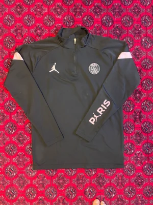 Svart PSG x Jordan träningsset - Säljer ett svart träningsset från PSG x Jordan med vita och rosa detaljer. Setet består av en långärmad tröja med halv dragkedja och matchande byxor med resår i midjan. Båda plaggen har PSG och Jumpman-loggor samt texten PARIS på ärm och ben.