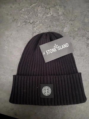 Svart ribbad mössa från Stone Island - Snygg svart ribbad mössa från Stone Island med klassisk logopatch framtill. Mössan har uppvikt kant och är perfekt för dig som vill ha en stilren accessoar med streetkänsla. Passar till de flesta outfits och håller dig varm under kyliga dagar.🙌 Helt oanvänd! Nypris 2000kr, mitt pris 200!😃