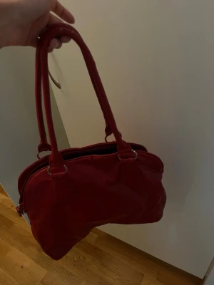 Röd handväska - Snygg röd handväska med silverfärgade metalldetaljer. Väskan har en mjuk, rundad form och dragkedja upptill. Perfekt för dig som vill ha en färgklick till din outfit. Finns ett litet fack med dragkedja. Några små sprickor finns men inget man tänker på (se bilder). 
