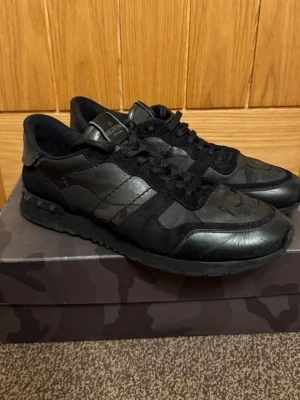 Svarta sneakers från Valentino Garavani - Snygga svarta sneakers från Valentino Garavani med coola kamouflagemönster och detaljer i både mocka och läder. Skorna har svarta snören, låg profil och en robust sula med nitar på hälen. Perfekta för dig som gillar exklusiv streetstyle.