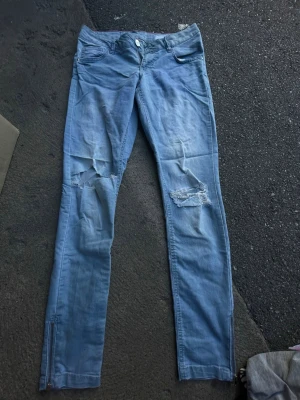 Ljusblå slitna skinny jeans - Säljer ett par ljusblå skinny jeans med slitningar och hål på båda knäna. Jeansen har dragkedjor vid bensluten och klassisk femficksdesign. Perfekta för en avslappnad och trendig look.