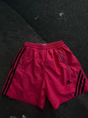 Rosa Adidas shorts med svarta ränder - Snygga rosa Adidas shorts med klassiska svarta ränder på sidorna och Adidas-logga framtill. Shortsen har elastisk midja med snörning, meshfoder och dragkedjeförsedda fickor. Perfekta för träning eller chill dagar. Funkar också som badkläder. 