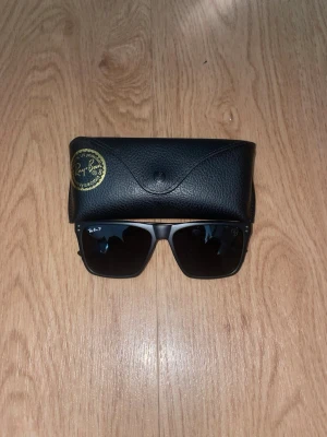 Svarta ferrari solglasögon från Ray-Ban - Snygga svarta solglasögon från Ray-Ban med klassisk fyrkantig form och mörka glas. Bågarna är i matt svart och har små silverdetaljer vid fronten. Medföljer ett svart fodral i skinnimitation. Perfekta för soliga dagar!