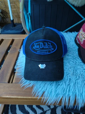 Svart och blå Von Dutch keps - Säljer en snygg truckerkeps från Von Dutch i svart och blått med broderad logga framtill. Kepsen har mesh på sidorna och bak för extra ventilation samt justerbar snapback. Klassisk böjd skärm och autenticitetsmärke på skärmen.