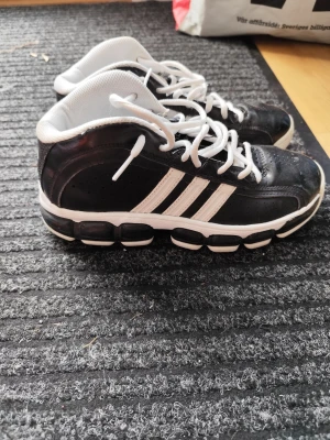 Svarta Adidas sneakers med vita detaljer - Snygga svarta sneakers från Adidas med klassiska vita ränder på sidorna och vit sula. Skorna har vit insida och vita skosnören. Ovandelen är i syntetmaterial och sulan är robust med tydlig dämpning. Perfekta för en sportig och trendig look.