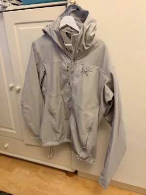 Ljusgrå vindjacka från Arc'teryx - Snygg ljusgrå vindjacka från Arc'teryx med huva och dragkedja framtill. Jackan har två sidofickor med dragkedja och justerbar nederkant. Tillverkad i ett lätt och smidigt material som passar perfekt för blåsiga dagar. Broderad logga på bröstet.