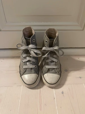 Grå höga Converse sneakers - Klassiska höga Converse sneakers i grå canvas med vita gummisulor och tåhättor. Skorna har silverfärgade öljetter och vita skosnören. Den ikoniska designen gör dem perfekta för en avslappnad och cool stil.