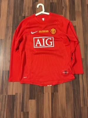 Manchester United långärmad retro tröja 2007-8 - Röd långärmad Manchester United matchtröja från Nike med AIG som sponsortryck och klubbmärke på bröstet. Tröjan har vita detaljer och texten 'FINAL MOSCOW 2008' broderad ovanför klubbmärket. Tillverkad i funktionsmaterial, perfekt för fotboll.