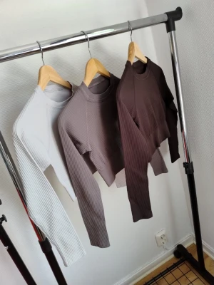 Ribbstickade crop tops i neutrala färger - Säljer tre ribbstickade crop tops med lång ärm och rund hals. Färgerna är vit, taupe och mörkbrun. Materialet är stretchigt och mjukt, perfekt för att skapa en clean och stilren look. Passar dig som gillar minimalistisk stil och vill ha något enkelt men snyggt.