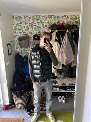 Moncler väst  - Snygg svart dunväst från Moncler med hög krage och blanka paneler. Västen har två dragkedjefickor framtill och Moncler-logga på bröstet. Perfekt för lager-på-lager och ger en trendig look. Stängs med både knappar och dragkedja.