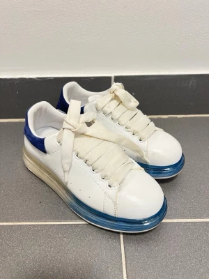 Vita Alexander mcqueen  - Snygga vita sneakers med breda vita skosnören, blå detaljer på hälen och en transparent blå sula. Skorna har en chunky design och är tillverkade i läder med textil på insidan. Perfekta för dig som gillar statement-sneakers med unik look.