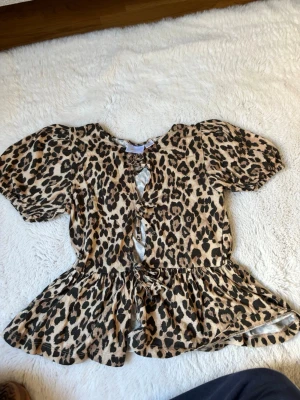 Leopardmönstrad topp från Gina Tricot - Säljer en leopardmönstrad topp från Gina Tricot med korta puffärmar och knyt framtill. Toppen har volang nedtill och är i mjukt material, perfekt för dig som vill sticka ut lite extra.