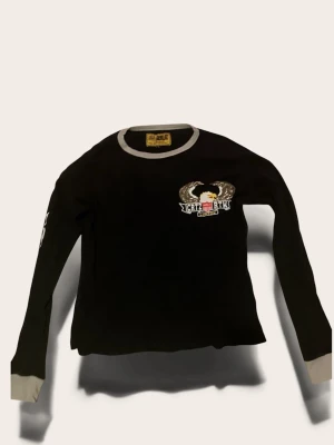 Svart långärmad topp från Corteiz - Cool svart långärmad topp från Corteiz med kontrasterande grå detaljer vid hals och ärmslut. Stort broderat örnmotiv och text på bröstet och ryggen, samt tryck på ena ärmen. Perfekt för dig som gillar streetwear och unika prints.