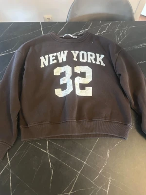 Brun sweatshirt med New York-tryck - Cool brun sweatshirt med vitt New York-tryck och siffran 32 på framsidan. Tröjan har rund hals och ribbade muddar vid ärmslut och nederkant. Perfekt för dig som gillar sportiga och avslappnade plagg. Materialet känns mjukt och bekvämt.