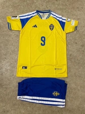 Sverige fotbollströja Adidas #9 - Gul och blå Sverige fotbollströja från Adidas med nummer 9 och namnet ISAK på ryggen. Tröjan har korta ärmar, blå detaljer och det svenska landslagets emblem på bröstet. Perfekt för fotbollsfans som vill visa sitt stöd på matchen.