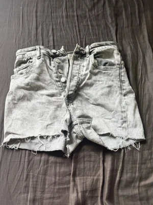 Gråa högmidjade jeansshorts - Säljer ett par gråa jeansshorts med fransiga kanter och klassisk femficksdesign. Shortsen har knäppning framtill och bälteshällor. Perfekta för en avslappnad stil.