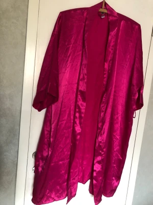 Morgonrock i ceriserosa satin - Snygg och glansig morgonrock/kimmono i ceriserosa satin med lös passform och trekvartsärmar. Perfekt för att chilla hemma i och ger en lyxig känsla tack vare det silkeslena materialet. Enkel att slänga på sig över pyjamasen.