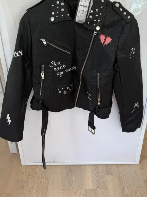 Svart skinnjacka med nitar och tryck - Cool svart skinnjacka från New Yorker med nitar, bältdetalj och dragkedjor. Framsidan har vita och röda tryck som 'You rock my world' och ett hjärta, medan ryggen har stort tryck 'HARD TO BREAK' och ett trasigt hjärta. Jackan har flera detaljer som blixtar, korsstygn och stjärnor.Oginal pris 700