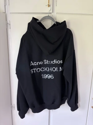 Svart hoodie Acne Studios Stockholm - Svart oversized hoodie från Acne Studios med stor huva och tryck på ryggen där det står 'Acne Studios STOCKHOLM 1996'. Hoodien har ribbade muddar vid ärmslut och nederkant. Perfekt för en avslappnad och trendig look. Köpt på Vinted så osäker på äktheten. Pris kan diskuteras vid snabb affär.
