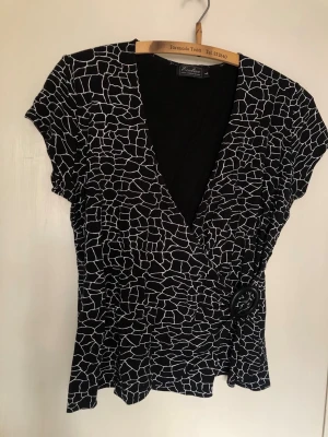 Svartvit omlottopp med bälte - Snygg svartvit omlottopp från Bon'A Parte med abstrakt mönster och markerad midja med rundt spänne. Djup v-ringning och korta ärmar ger en trendig look. Perfekt till jeans eller kjol för en cool outfit.