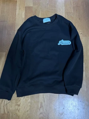 Svart sweatshirt Casablanca Aiir - Svart sweatshirt från Casablanca med trycket 'Aiir' på bröstet och stort färgglatt flygplansmotiv samt Casablanca-logga på ryggen. Tröjan har rund hals, ribbade muddar och är gjord i mjukt bomullsmaterial. Perfekt för en streetwear-look. 