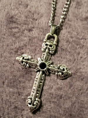Kedjehalsband med kors med svart sten - Snyggt halsband i rostfritt stål med ett detaljerat korshänge silverfärgat. Korset har dekorativa mönster och en svart sten i mitten som ger en cool kontrast. Kedjan är av klassisk modell och passar perfekt till streetwear eller rockig stil.