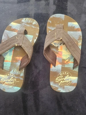 Warp flip-flops med palmtryck - Snygga flip-flops från Warp med breda bruna textilremmar och färgglad sula med palm- och strandmotiv. Perfekta för stranden eller sommardagar. Lätta att ta på och av, med mjuk fotbädd och slitstark gummisula.