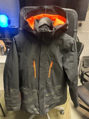 Svart McKinley skidjacka 158 med orange detaljer och skidbyxor 152 - Snygg svart skidjacka 158 med tillhörande skidbyxor 152 från McKinley med orange foder och dragkedjor. Jackan har huva, två framfickor med dragkedja och diskret camomönster nedtill. Perfekt för dig som vill ha en sportig och cool look. Materialet är slitstarkt och vattenavvisande. Går att köpa för sig om önskas då jacka 450:-  byxa 350:-. Kanonskick 