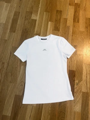 Vit t-shirt från J.Lindeberg - Klassisk vit t-shirt från J.Lindeberg med diskret logga på bröstet. T-shirten har rund halsringning och korta ärmar. Tillverkad i mjuk bomull som känns skön mot huden. Perfekt basplagg till garderoben.