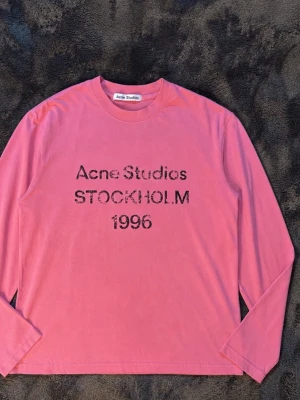 Rosa långärmad topp Acne Studios - Rosa långärmad topp från Acne Studios med tryck 'Acne Studios STOCKHOLM 1996' framtill. Klassisk rund halsringning och relaxed fit. Perfekt för dig som gillar streetwear och vill ha något stilrent men ändå statement. Materialet känns mjukt och skönt mot huden. Storlek M men passar S också.