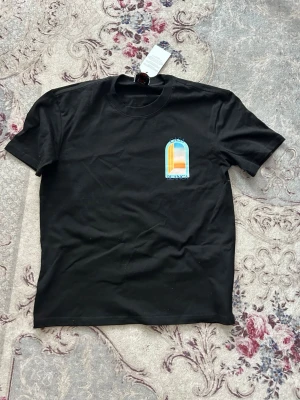 Svart Casa Blanca t-shirt - Snygg svart t-shirt från Casa Blanca med färgglad grafisk print på bröstet i blått, orange och turkost. Klassisk rund hals och korta ärmar. Perfekt för dig som vill ha en stilren men ändå unik look. Lite bred runt midjan men ger en snygg baggy look. 