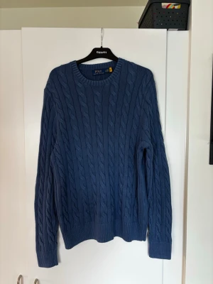 Blå kabelstickad tröja Polo Ralph Lauren - Kabelstickad blå tröja från Polo Ralph Lauren med rund halsringning och diskret broderad logga på bröstet. I mycket bra skick, endast provat den. 