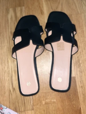 Svarta slip-in sandaler med fyrkantig tå - Stilrena svarta slip-in sandaler med breda remmar och fyrkantig tå. Sandalerna har öppen häl och ljus innersula, samt en modern design med utskurna detaljer på ovandelen. Perfekta för en trendig och avslappnad look.