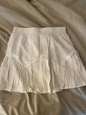 Vit kortkjol - Söt vit kortkjol med volang och dekorativ spetsdetalj i blommönster. Kjolen har en elastisk midja och är gjord i ett lätt, luftigt material som passar perfekt till varma dagar. Enkel att matcha med olika toppar för en fräsch look.