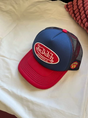 Von Dutch röd och blå truckerkeps - Snygg truckerkeps från Von Dutch med blå framsida, röd skärm och röd knapp på toppen. Kepsen har svart mesh baktill och broderad Von Dutch-logga framtill. 