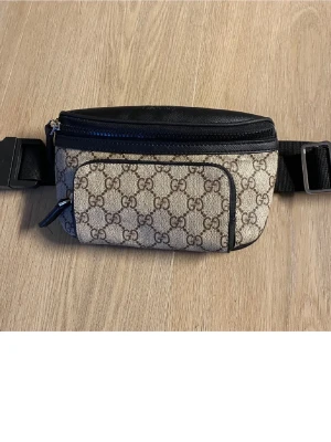 Gucci beige axelväska med GG-mönster - Snygg axelväska från Gucci i beige canvas med klassiskt GG-mönster och svarta läderdetaljer. Väskan har flera fack med dragkedja och justerbar rem för perfekt passform. Perfekt för dig som vill ha både stil och funktion.